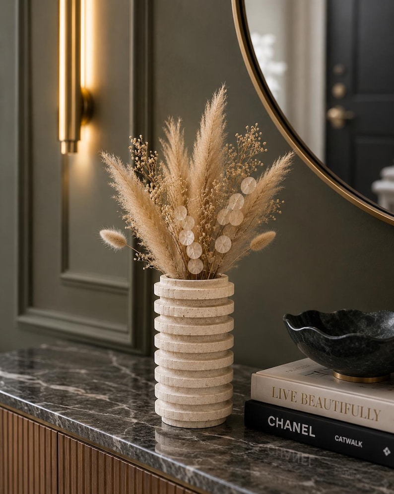 May include: A cylindrical beige stone vase with a ribbed, stacked-ring texture holds a dried floral arrangement of tan pampas grass, fluffy bunny tails, and small white flowers with circular accents, creating a neutral-toned decorative display.