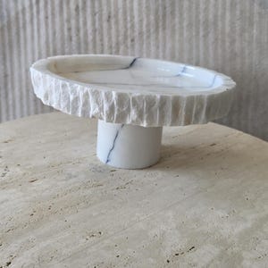 May include: A white marble pedestal bowl featuring a smooth, polished interior surface with subtle grey veining and a contrasting rough, chiseled exterior edge. The bowl is supported by a cylindrical marble base that matches the bowl's white color.