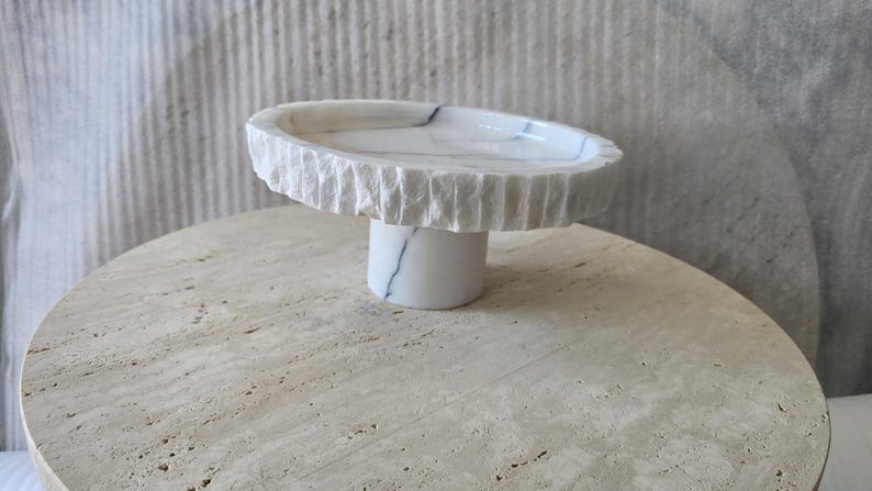 May include: A white marble pedestal bowl featuring a smooth, polished interior surface with subtle grey veining and a contrasting rough, chiseled exterior edge. The bowl is supported by a cylindrical marble base that matches the bowl's white color.