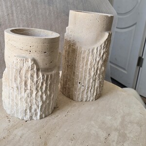 May include: Two cylindrical beige stone vessels with a porous, natural texture. Each piece features a smooth, circular top opening and a rough, vertically grooved exterior surface that resembles tree bark, crafted from light-colored travertine stone.