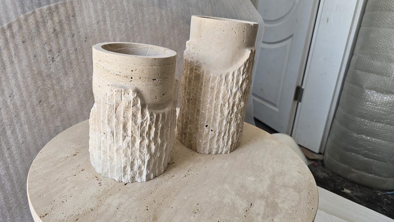 May include: Two cylindrical beige stone vessels with a porous, natural texture. Each piece features a smooth, circular top opening and a rough, vertically grooved exterior surface that resembles tree bark, crafted from light-colored travertine stone.