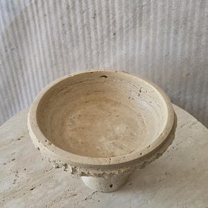 K&ouml;nnte beinhalten: Eine beigefarbene Schale aus Travertin mit kreisf&ouml;rmiger Form und einer por&ouml;sen, strukturierten Oberfl&auml;che. Die Schale verf&uuml;gt &uuml;ber einen breiten, flachen Rand und einen konischen Fu&szlig;, der die nat&uuml;rlichen Maserungen und Erdt&ouml;ne des Steins zeigt.
