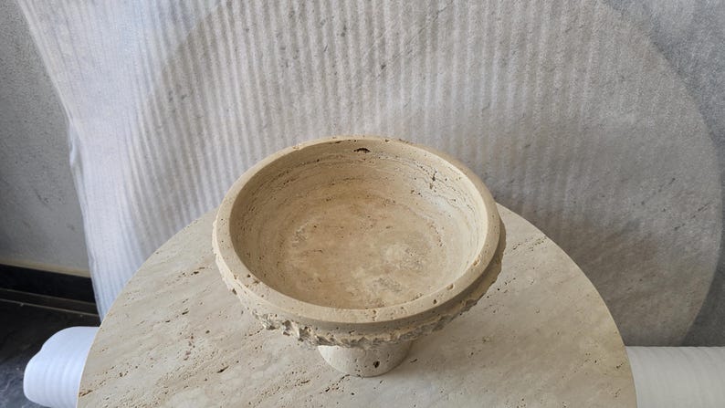 K&ouml;nnte beinhalten: Eine beigefarbene Schale aus Travertin mit kreisf&ouml;rmiger Form und einer por&ouml;sen, strukturierten Oberfl&auml;che. Die Schale verf&uuml;gt &uuml;ber einen breiten, flachen Rand und einen konischen Fu&szlig;, der die nat&uuml;rlichen Maserungen und Erdt&ouml;ne des Steins zeigt.