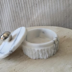 May include: A round white marble container with grey veining and a textured exterior. The matching lid features a smooth, polished silver-toned spherical handle. The container is open, revealing a smooth interior basin for storage or display.