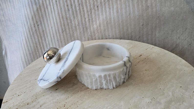 May include: A round white marble container with grey veining and a textured exterior. The matching lid features a smooth, polished silver-toned spherical handle. The container is open, revealing a smooth interior basin for storage or display.