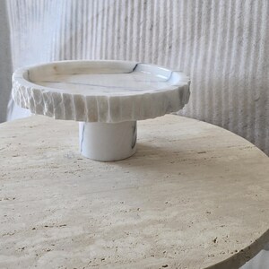 May include: A white marble cake stand featuring a circular top with a textured, chiseled edge and a smooth, cylindrical base. The stone displays subtle grey veining throughout the surface, providing a natural contrast between the rough rim and the polished pedestal.