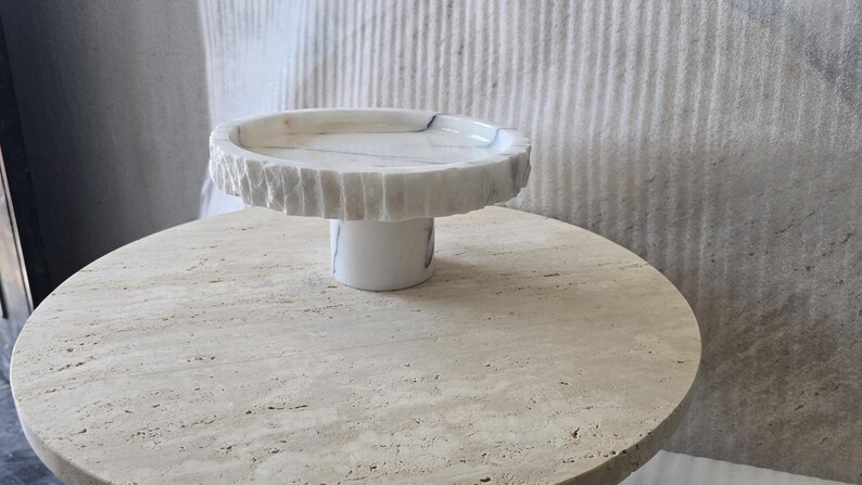 May include: A white marble cake stand featuring a circular top with a textured, chiseled edge and a smooth, cylindrical base. The stone displays subtle grey veining throughout the surface, providing a natural contrast between the rough rim and the polished pedestal.