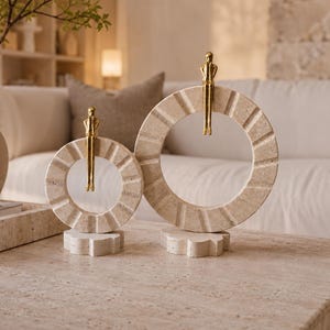 Pu&ograve; includere: Due sculture decorative composte da anelli circolari in pietra beige con scanalature radiali testurizzate. Ogni anello poggia su una base a forma di fiore ed &egrave; sormontato da una piccola figura metallica color oro seduta sul bordo superiore.
