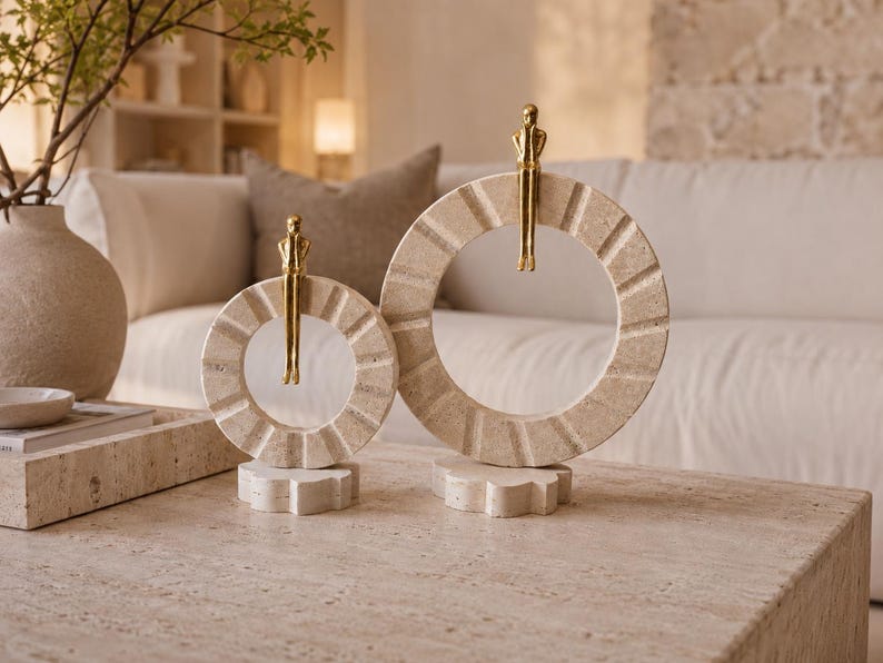 Pu&ograve; includere: Due sculture decorative composte da anelli circolari in pietra beige con scanalature radiali testurizzate. Ogni anello poggia su una base a forma di fiore ed &egrave; sormontato da una piccola figura metallica color oro seduta sul bordo superiore.