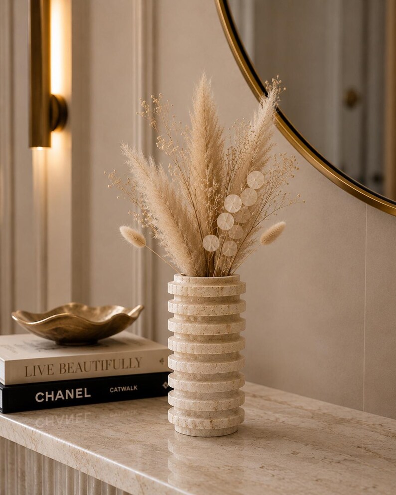 May include: A cylindrical beige stone vase with a ribbed, stacked-ring texture. The vase is filled with a dried floral arrangement consisting of tall, feathery pampas grass, delicate baby's breath, and small, soft, round bunny tail grass stems.