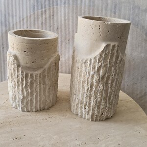 May include: Two cylindrical beige travertine stone vessels with a porous, natural texture. The lower sections feature a vertical, rough-hewn chiseled pattern, while the upper rims are smooth and circular. The vessels vary in height.