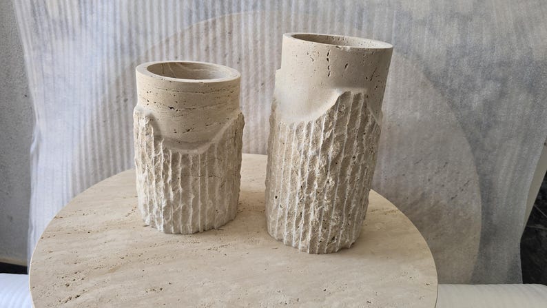 May include: Two cylindrical beige travertine stone vessels with a porous, natural texture. The lower sections feature a vertical, rough-hewn chiseled pattern, while the upper rims are smooth and circular. The vessels vary in height.