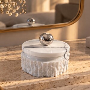 May include: A round white marble box with a textured, rough-hewn exterior and a smooth, polished lid. The lid features a central, spherical, silver-toned metal knob with a hammered finish and dark grey veining running across the white stone surface.