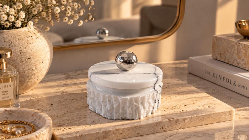 May include: A round white marble box with a textured, rough-hewn exterior and a smooth, polished lid. The lid features a central, spherical, silver-toned metal knob with a hammered finish and dark grey veining running across the white stone surface.