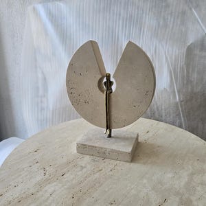 K&ouml;nnte beinhalten: Eine Skulptur aus beigem Travertin mit einer kreisf&ouml;rmigen Steinscheibe und einem keilf&ouml;rmigen Ausschnitt. Eine schlanke, metallisch bronzefarbene Figur sitzt in der zentralen &Ouml;ffnung, wobei die Beine &uuml;ber den Steinsockel h&auml;ngen.