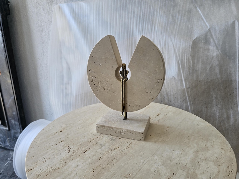 K&ouml;nnte beinhalten: Eine Skulptur aus beigem Travertin mit einer kreisf&ouml;rmigen Steinscheibe und einem keilf&ouml;rmigen Ausschnitt. Eine schlanke, metallisch bronzefarbene Figur sitzt in der zentralen &Ouml;ffnung, wobei die Beine &uuml;ber den Steinsockel h&auml;ngen.