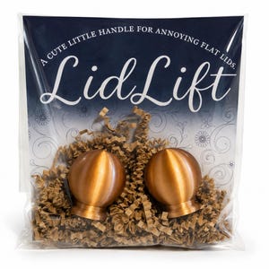 May include: Two copper-coloured spherical handles with flat bases packaged in a clear plastic bag with brown paper filler. The packaging features the text "LidLift" and "A CUTE LITTLE HANDLE FOR ANNOYING FLAT LIDS" printed in white on a dark blue background.