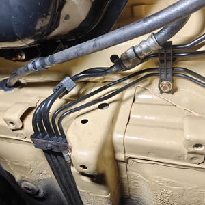 May include: A beige painted automotive chassis section featuring a series of thin black metal brake lines routed through a black plastic mounting bracket. A flexible rubber hose connects to the metal lines, secured by a bolt and a plastic clip.