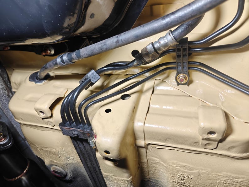 May include: A beige painted automotive chassis section featuring a series of thin black metal brake lines routed through a black plastic mounting bracket. A flexible rubber hose connects to the metal lines, secured by a bolt and a plastic clip.