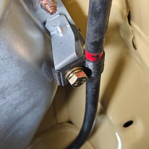 May include: A silver metal bracket secures a black rubber hose against a light tan surface. The assembly features a gold-colored hexagonal bolt with visible markings, a rusted nut on the upper mounting point, and a red band on the hose.