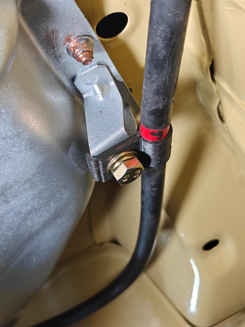 May include: A silver metal bracket secures a black rubber hose against a light tan surface. The assembly features a gold-colored hexagonal bolt with visible markings, a rusted nut on the upper mounting point, and a red band on the hose.