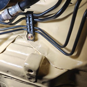 May include: A series of black metal automotive brake lines secured by a black plastic mounting bracket with a gold-colored nut and bolt, all installed against a smooth, light tan painted metal chassis surface within an engine compartment.