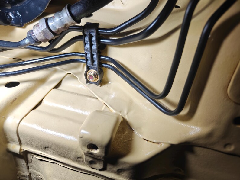 May include: A series of black metal automotive brake lines secured by a black plastic mounting bracket with a gold-colored nut and bolt, all installed against a smooth, light tan painted metal chassis surface within an engine compartment.