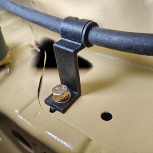 May include: A black metal bracket secured with a silver-colored hexagonal bolt and washer, holding a black rubber hose in place against a smooth, tan-colored painted surface. The bracket features a curved top section for the hose.