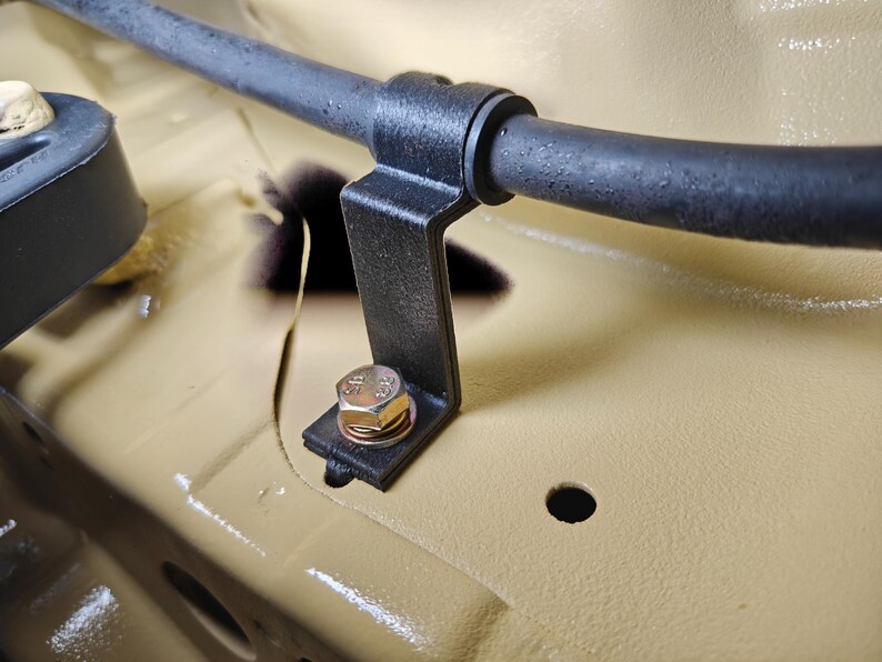 May include: A black metal bracket secured with a silver-colored hexagonal bolt and washer, holding a black rubber hose in place against a smooth, tan-colored painted surface. The bracket features a curved top section for the hose.