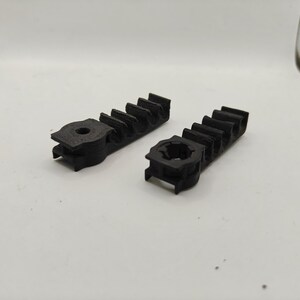 May include: Two black, 3D-printed plastic components featuring a linear gear rack design with multiple teeth and a circular mounting base at one end. Each piece has a textured surface and a central hole for attachment, showing a functional mechanical part.