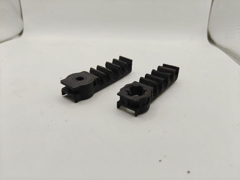 May include: Two black, 3D-printed plastic components featuring a linear gear rack design with multiple teeth and a circular mounting base at one end. Each piece has a textured surface and a central hole for attachment, showing a functional mechanical part.