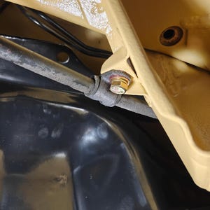 May include: A close-up view of a gold-colored metal bolt securing a dark gray cylindrical rod to a tan-painted metal frame. The assembly is positioned above a smooth, glossy black surface, with visible black wiring tucked behind the frame structure.