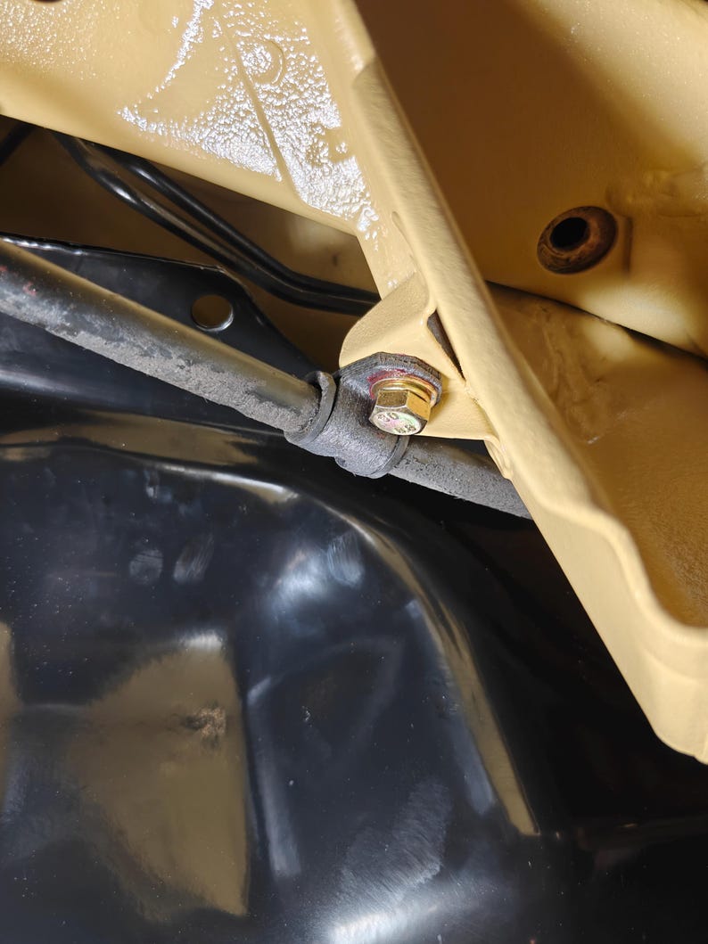 May include: A close-up view of a gold-colored metal bolt securing a dark gray cylindrical rod to a tan-painted metal frame. The assembly is positioned above a smooth, glossy black surface, with visible black wiring tucked behind the frame structure.