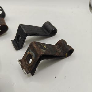 May include: Two L-shaped metal mounting clips with a circular hole at one end and a curved loop at the other. One clip is matte black with a textured finish, while the other is weathered, showing significant brown rust and metallic discoloration.