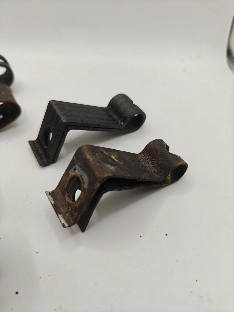 May include: Two L-shaped metal mounting clips with a circular hole at one end and a curved loop at the other. One clip is matte black with a textured finish, while the other is weathered, showing significant brown rust and metallic discoloration.