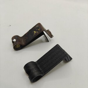 May include: Two L-shaped metal clips with a rounded hinge-like end. One clip is dark brown with visible signs of oxidation and wear, while the other is matte black with a textured finish. Both clips feature a bent design for mounting or securing.