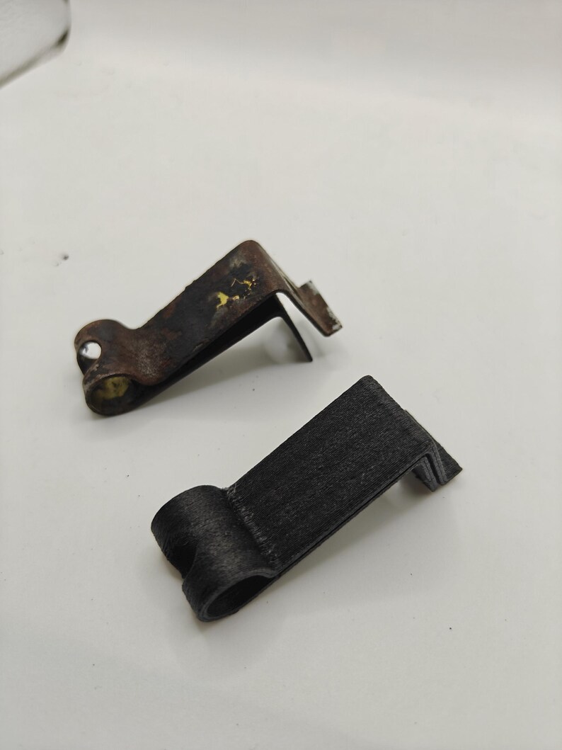 May include: Two L-shaped metal clips with a rounded hinge-like end. One clip is dark brown with visible signs of oxidation and wear, while the other is matte black with a textured finish. Both clips feature a bent design for mounting or securing.
