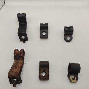 May include: A collection of six metal mounting brackets arranged in two rows. The top row features three black, clean brackets, while the bottom row contains three matching brackets showing significant rust and oxidation, each with a circular hole.