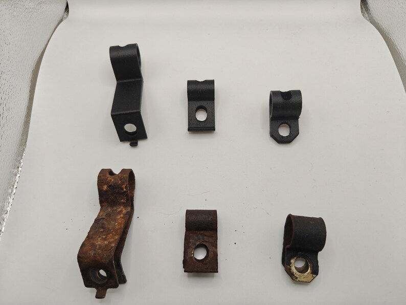 May include: A collection of six metal mounting brackets arranged in two rows. The top row features three black, clean brackets, while the bottom row contains three matching brackets showing significant rust and oxidation, each with a circular hole.