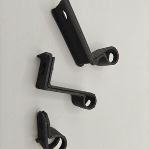 May include: Three black 3D-printed mounting brackets with a textured surface. Each bracket features an L-shaped profile with a flat mounting plate containing a circular hole and a curved, cylindrical hook section at the opposite end.