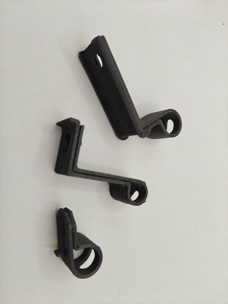May include: Three black 3D-printed mounting brackets with a textured surface. Each bracket features an L-shaped profile with a flat mounting plate containing a circular hole and a curved, cylindrical hook section at the opposite end.