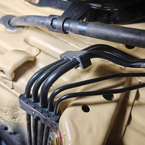 May include: A black plastic cable management clip securing four thin black hoses against a tan painted metal surface. The clip features a rectangular base with four parallel channels that hold the hoses in place, positioned near a larger black hose.