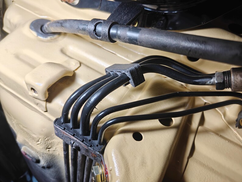 May include: A black plastic cable management clip securing four thin black hoses against a tan painted metal surface. The clip features a rectangular base with four parallel channels that hold the hoses in place, positioned near a larger black hose.
