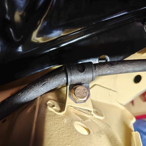 May include: A dark gray metal sway bar secured by a black rubber bushing and a gold-colored hexagonal bolt with the text 8.8 stamped on its head. The assembly is mounted to a beige-painted metal chassis component under a black panel.