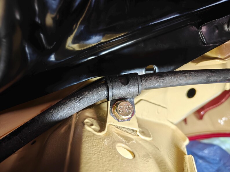 May include: A dark gray metal sway bar secured by a black rubber bushing and a gold-colored hexagonal bolt with the text 8.8 stamped on its head. The assembly is mounted to a beige-painted metal chassis component under a black panel.