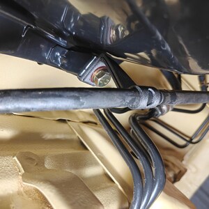 May include: A black automotive brake line assembly featuring a series of curved metal tubes and a flexible hose secured with a metal bracket and a hexagonal bolt, mounted against a light tan painted chassis panel within a vehicle undercarriage.