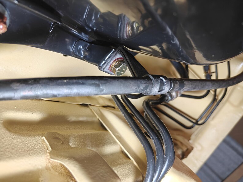 May include: A black automotive brake line assembly featuring a series of curved metal tubes and a flexible hose secured with a metal bracket and a hexagonal bolt, mounted against a light tan painted chassis panel within a vehicle undercarriage.