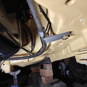 May include: The underside of a vehicle chassis painted in a light tan color, featuring a black metal bracket secured with a bolt, various black rubber hoses, and a metallic rod running horizontally across the frame near the engine components.