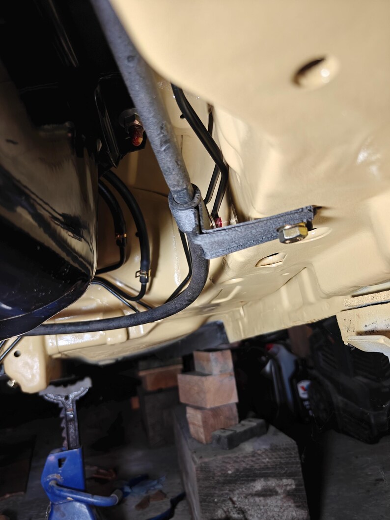 May include: The underside of a vehicle chassis painted in a light tan color, featuring a black metal bracket secured with a bolt, various black rubber hoses, and a metallic rod running horizontally across the frame near the engine components.