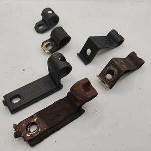 May include: A collection of six metal mounting brackets and clamps of varying sizes and conditions. Some pieces feature a smooth black finish, while others exhibit significant surface rust and oxidation, each with a single circular hole.
